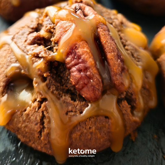 Close-up of a chocolate muffin with caramel drizzle and pecans on a dark surface, branded 'ketome'.